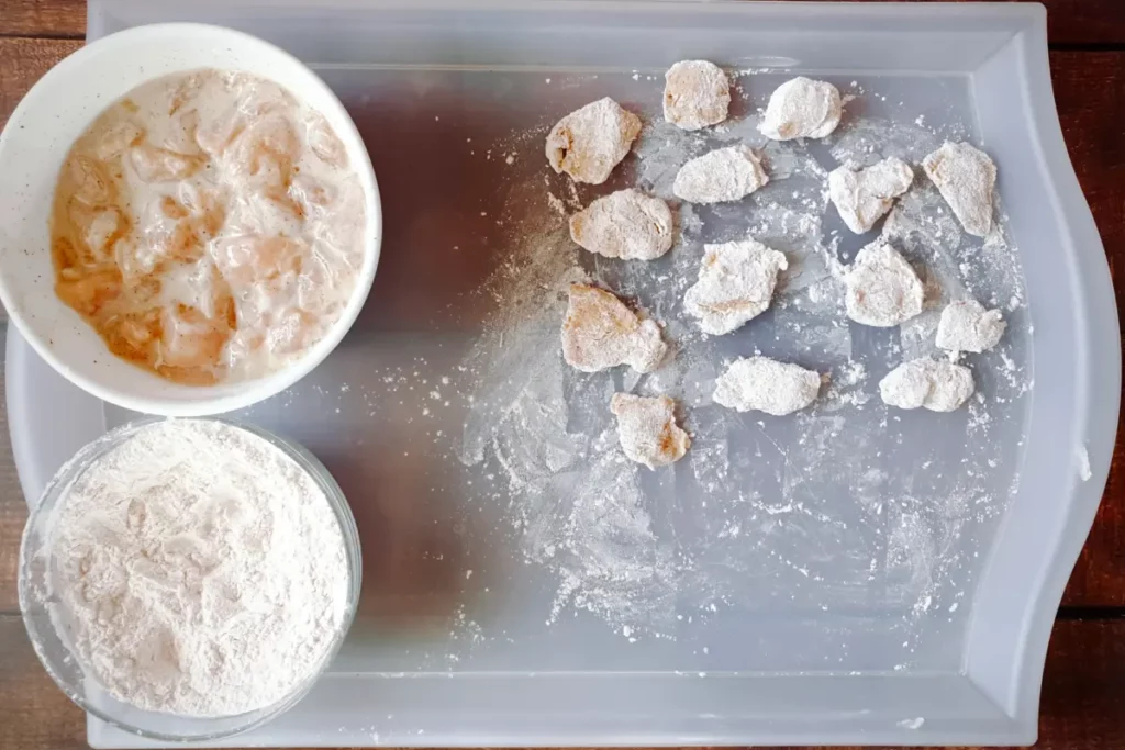 Two bowls of wet and dry ingredients on the left and a few dredged chicken pieces on the right, all over a tray.